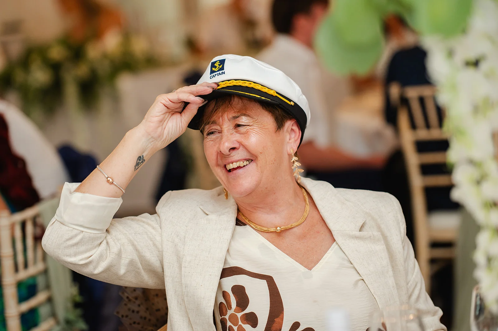 A smiling wedding guest wearing a captain hat and a cream blazer laughs heartily during the reception.