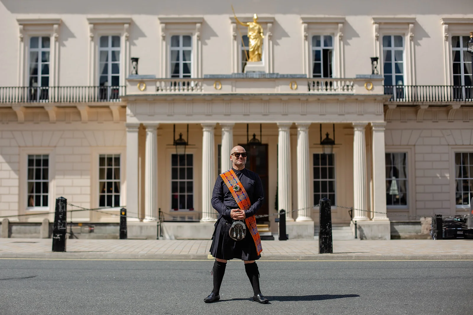 A groom in traditional highland dress and kilt stands proudly on a city street in front of a grand neoclassical building on a sunny day.
