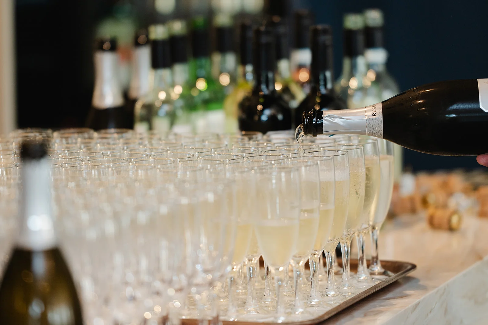 A person pours sparkling wine from a bottle into a large tray of champagne flutes arranged for a wedding drinks reception.
