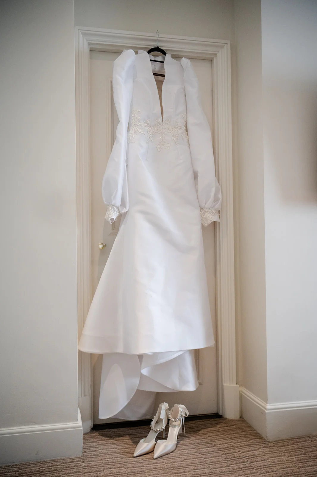 An elegant white long-sleeved satin wedding dress hangs neatly in a white doorway with a pair of embellished bridal heels placed on the carpet below.