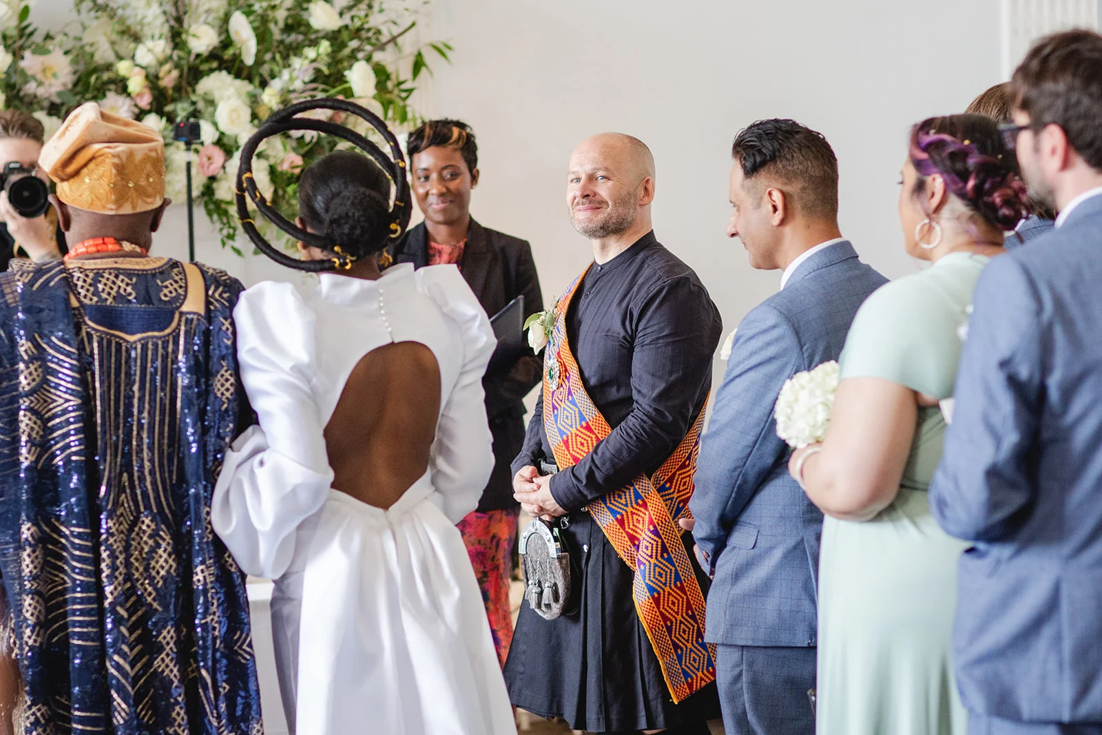 An intercultural wedding ceremony featuring a bride in a sculptural white gown with a dramatic circular headpiece and a groom in a black kilt with a colourful patterned sash.