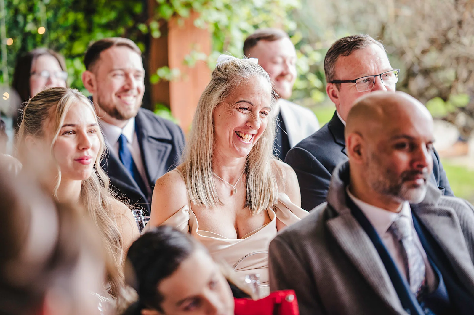 Wedding guests seated together laughing happily during a speech in a naturally lit outdoor setting.