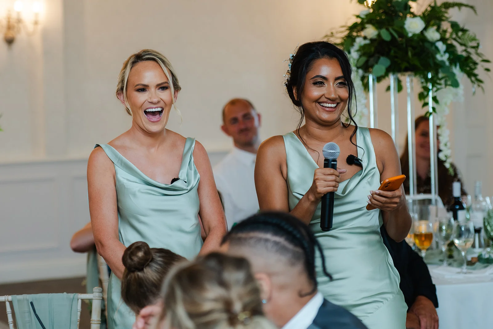 Two bridesmaids in sage green satin cowl neck dresses laugh joyfully while delivering a speech during the wedding reception.