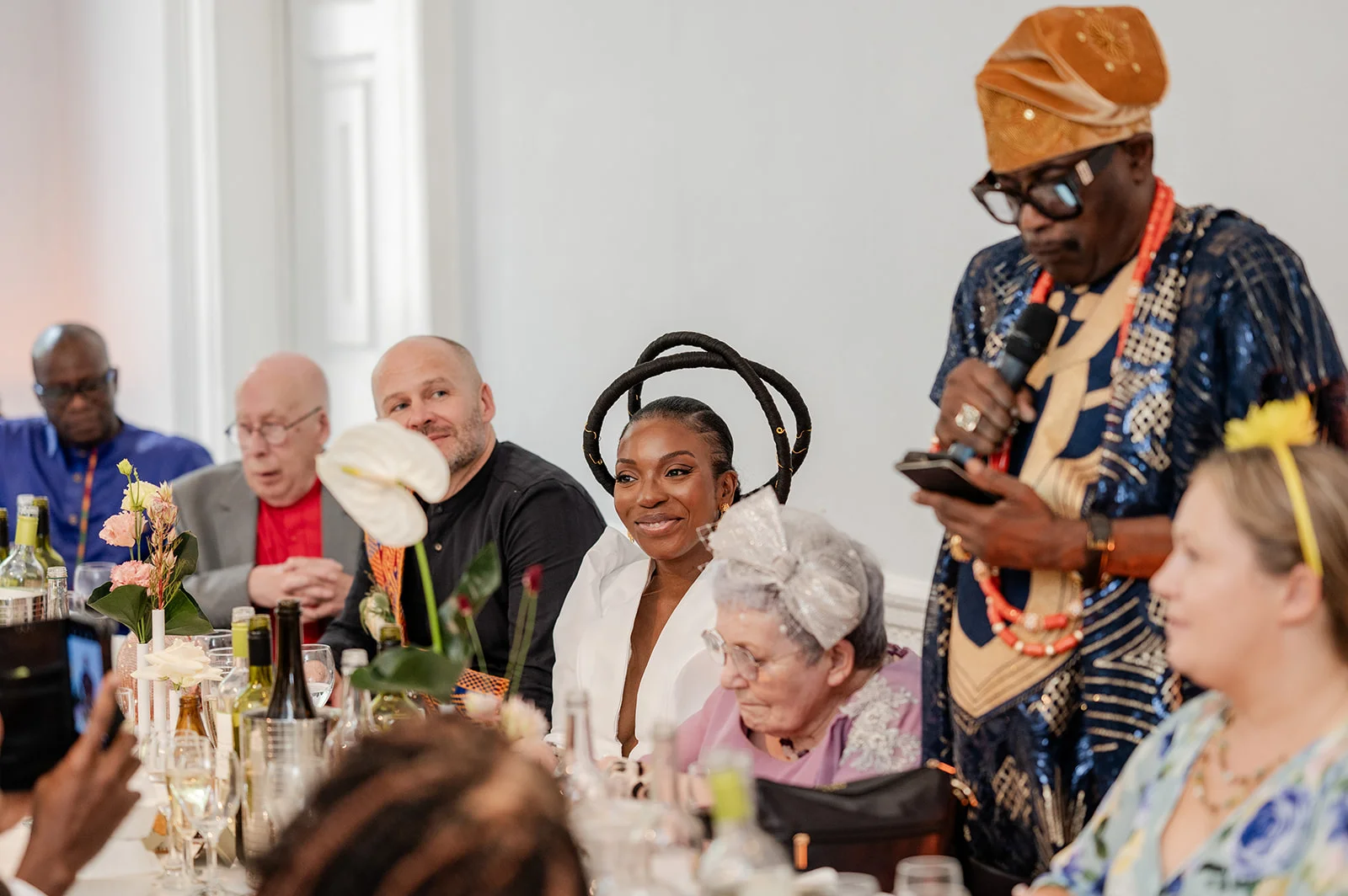 A guest wearing traditional Nigerian attire with a patterned tunic and cap speaks into a microphone during a wedding reception dinner while seated guests listen.