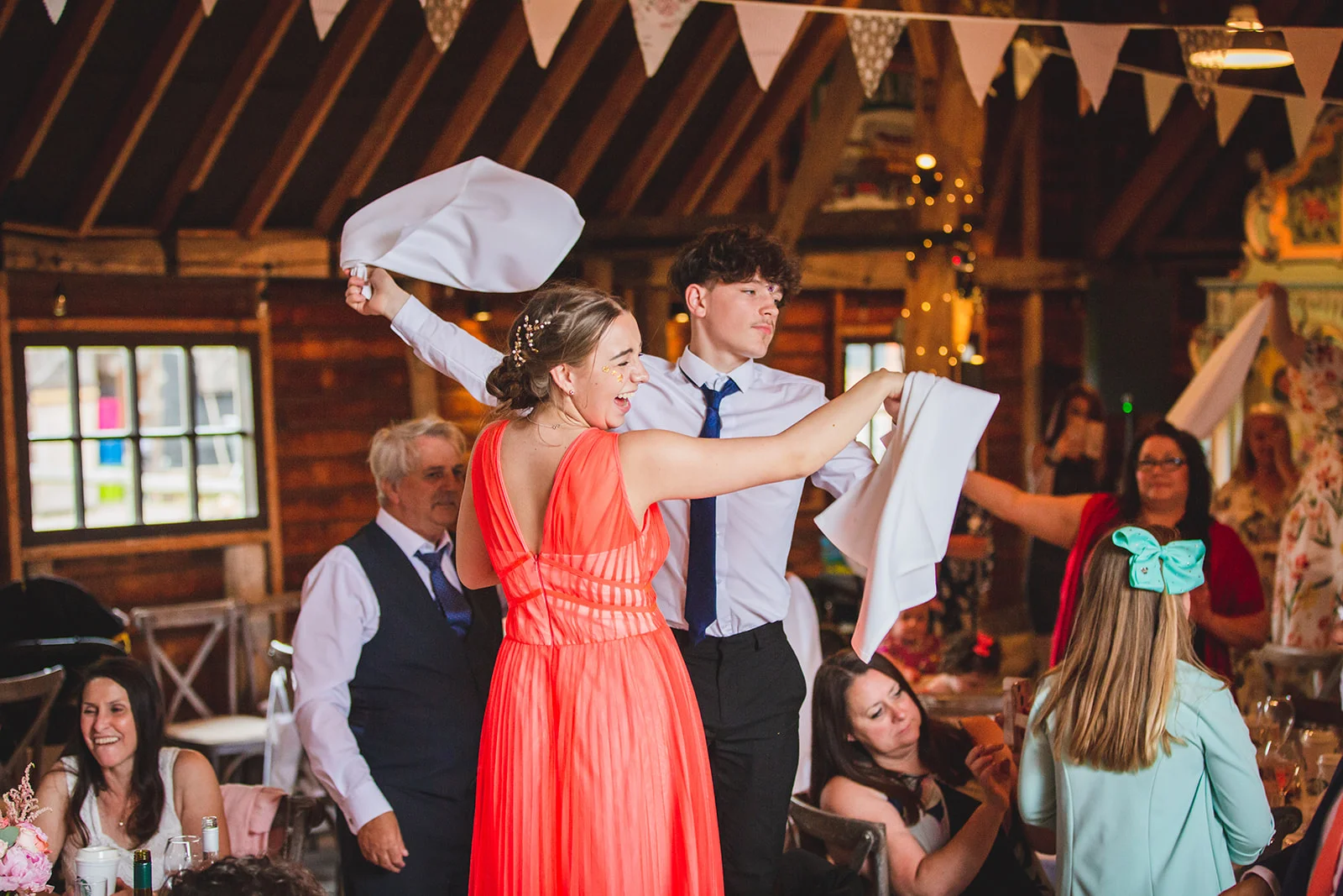 Wedding guests enthusiastically wave white napkins in the air during the wedding reception inside a rustic barn decorated with exposed wooden beams and bunting.