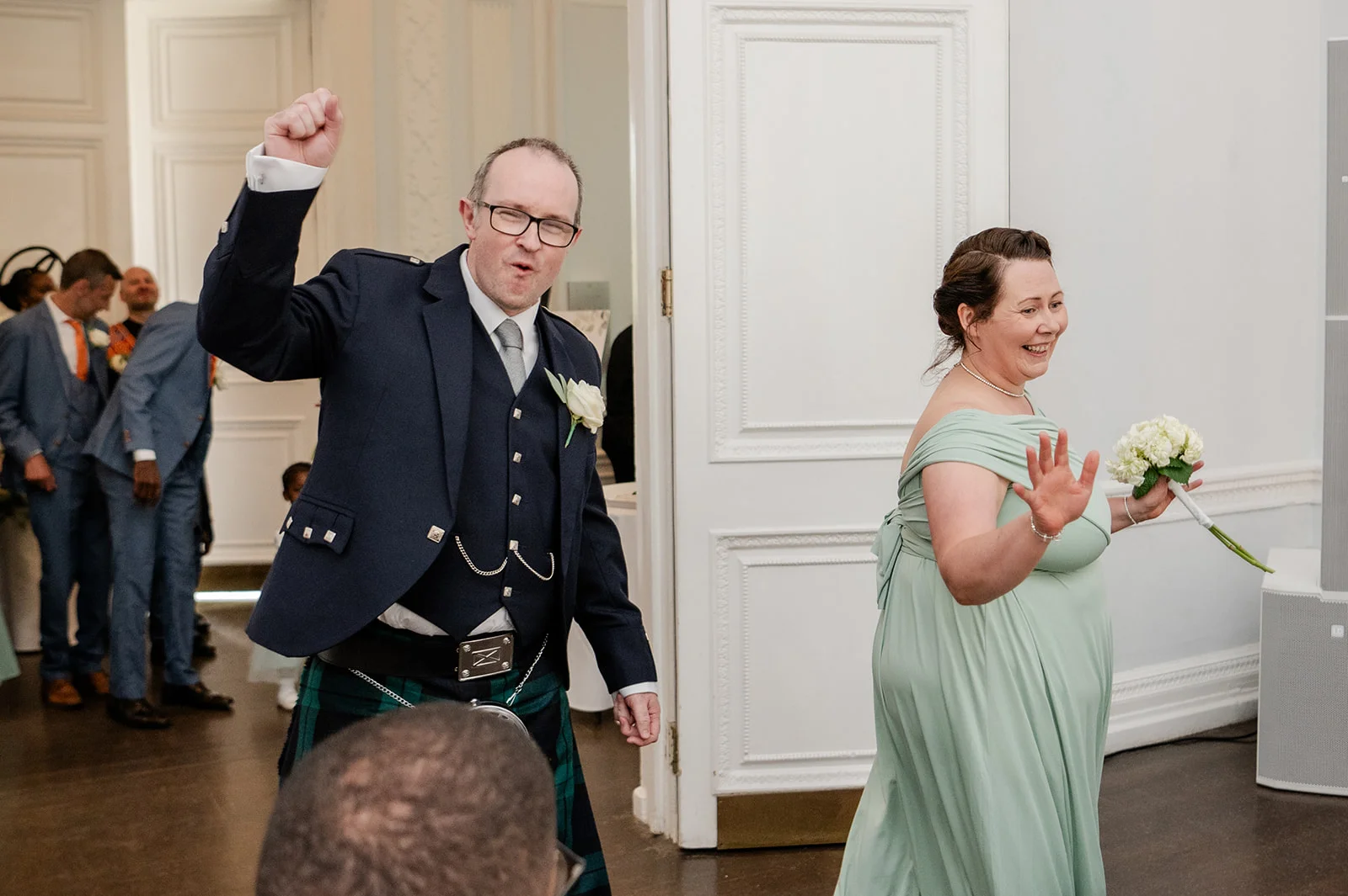 A joyful couple make their entrance at a wedding reception, with the groom raising his arm in celebration while wearing a traditional kilt and the bride smiling warmly in a sage green dress holding a white posy bouquet.