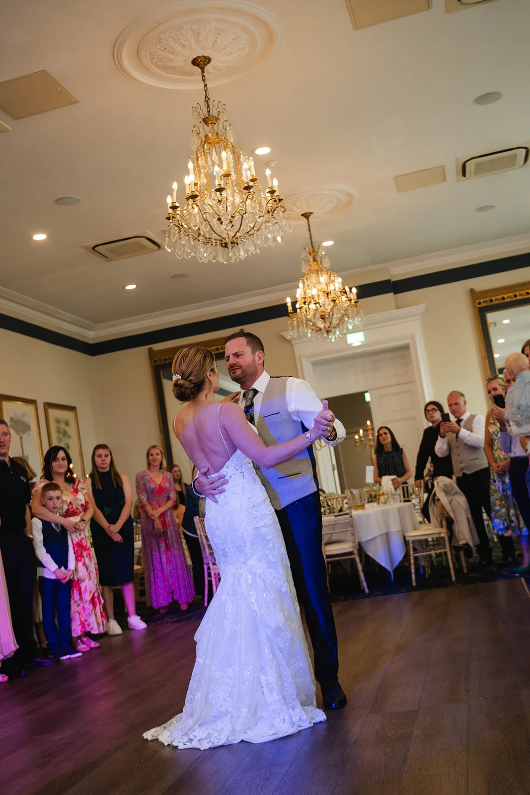 Newlyweds share their first dance in an elegant ballroom illuminated by crystal chandeliers, surrounded by watching guests.