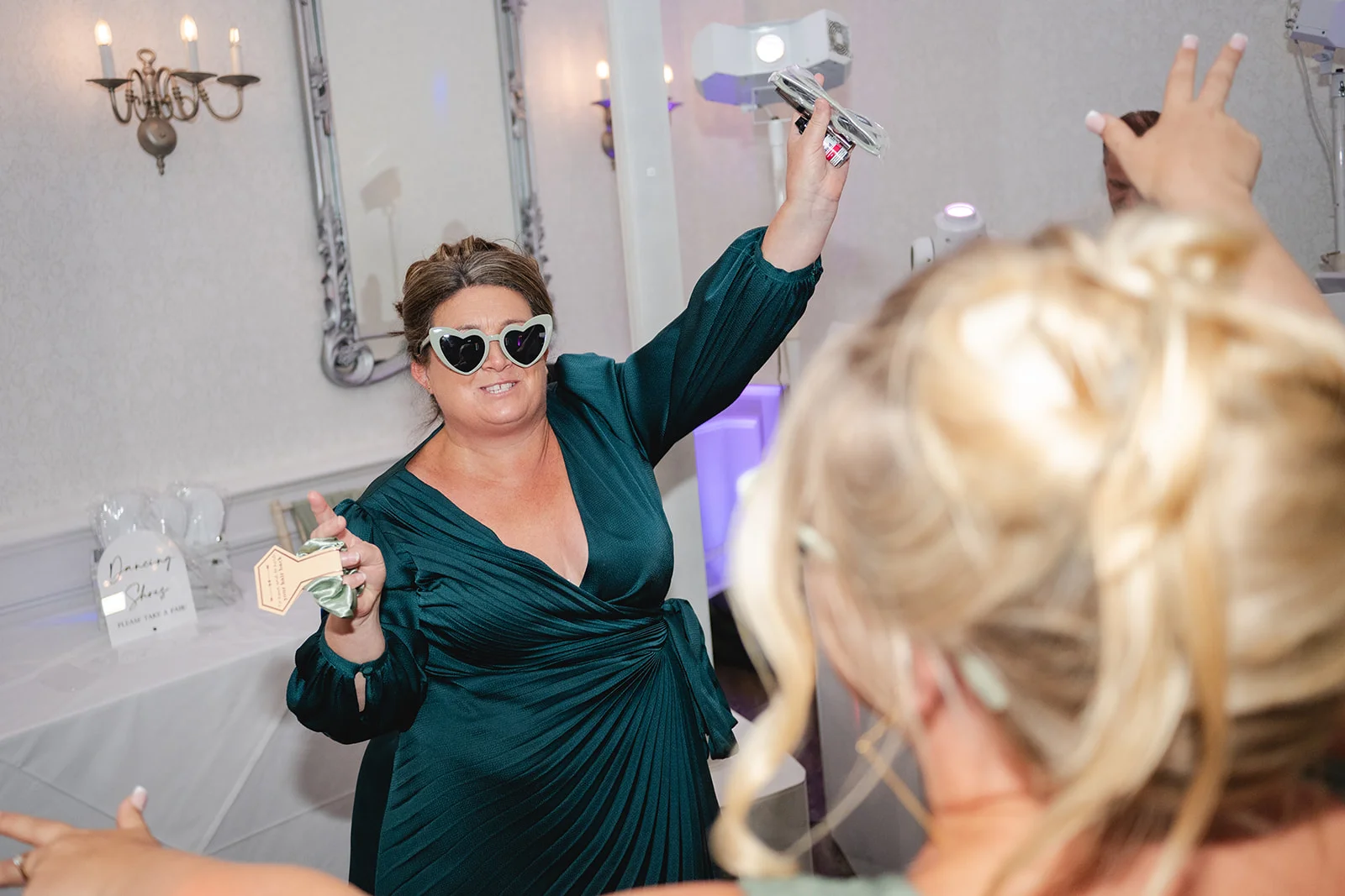 A joyful wedding guest wearing heart-shaped sunglasses and a dark green dress dances with her arms raised during the evening reception.