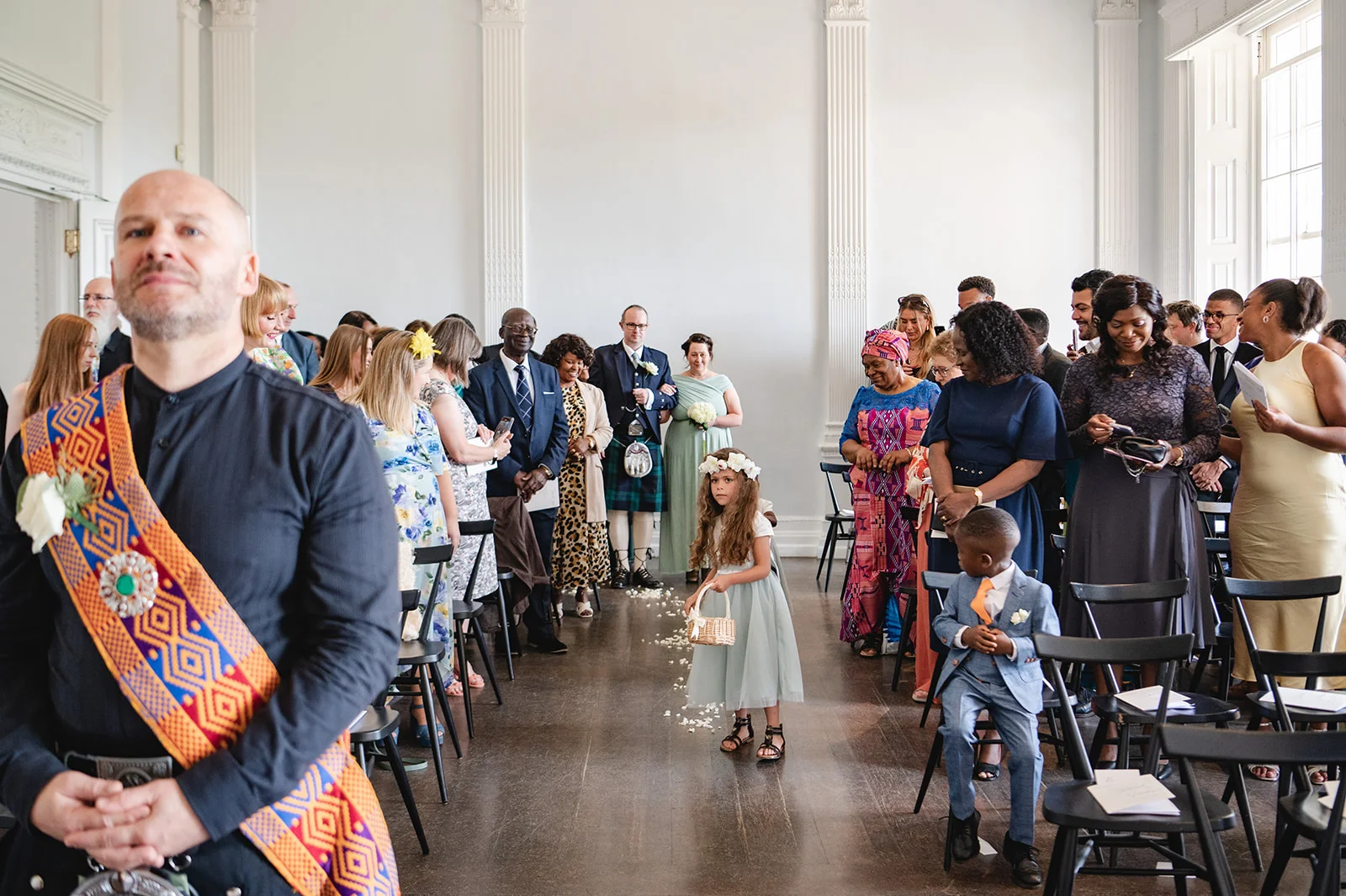 A young flower girl holding a small basket walks down the aisle during a bright indoor wedding ceremony attended by seated guests.