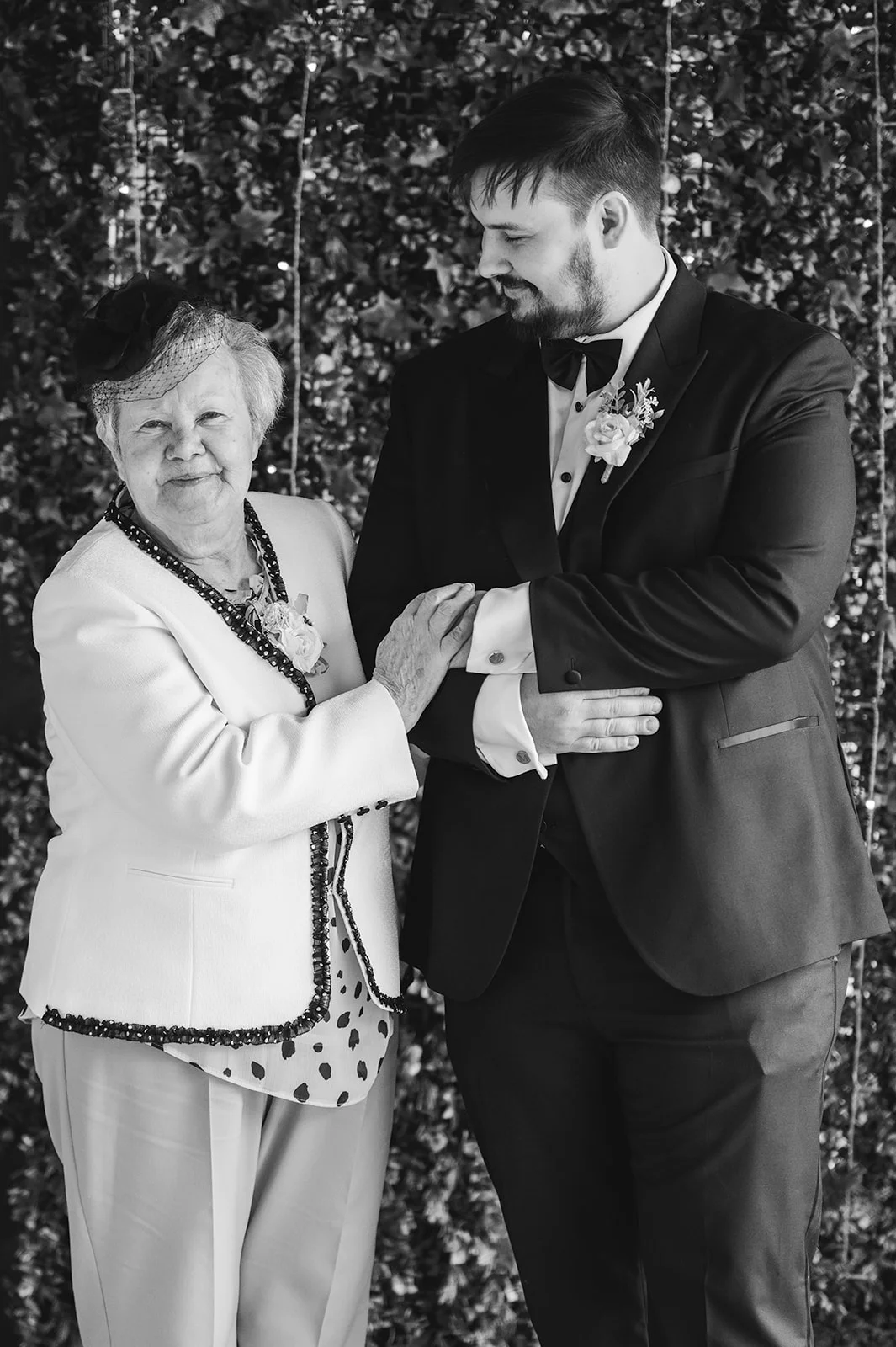 A smiling older woman wearing a fascinator and jacket stands beside a groom in a black tuxedo, gently holding his arm in front of a leafy wall.