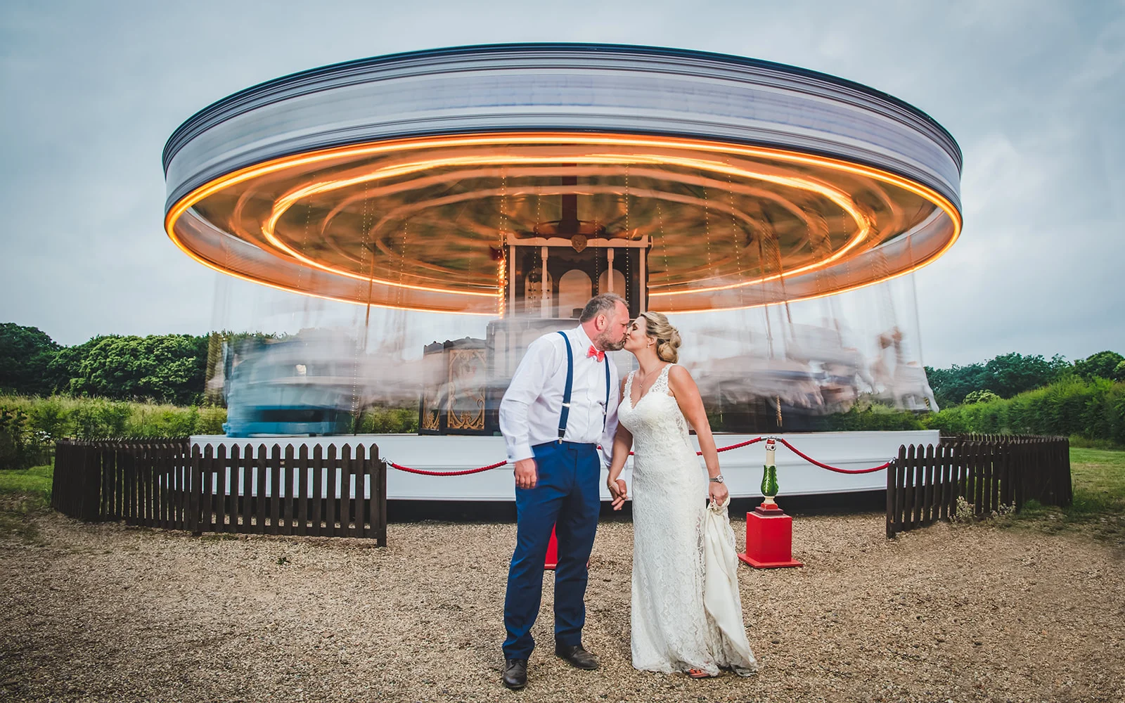 A newlywed couple shares a romantic kiss in front of a brightly lit vintage carousel with motion blur at an outdoor wedding reception.