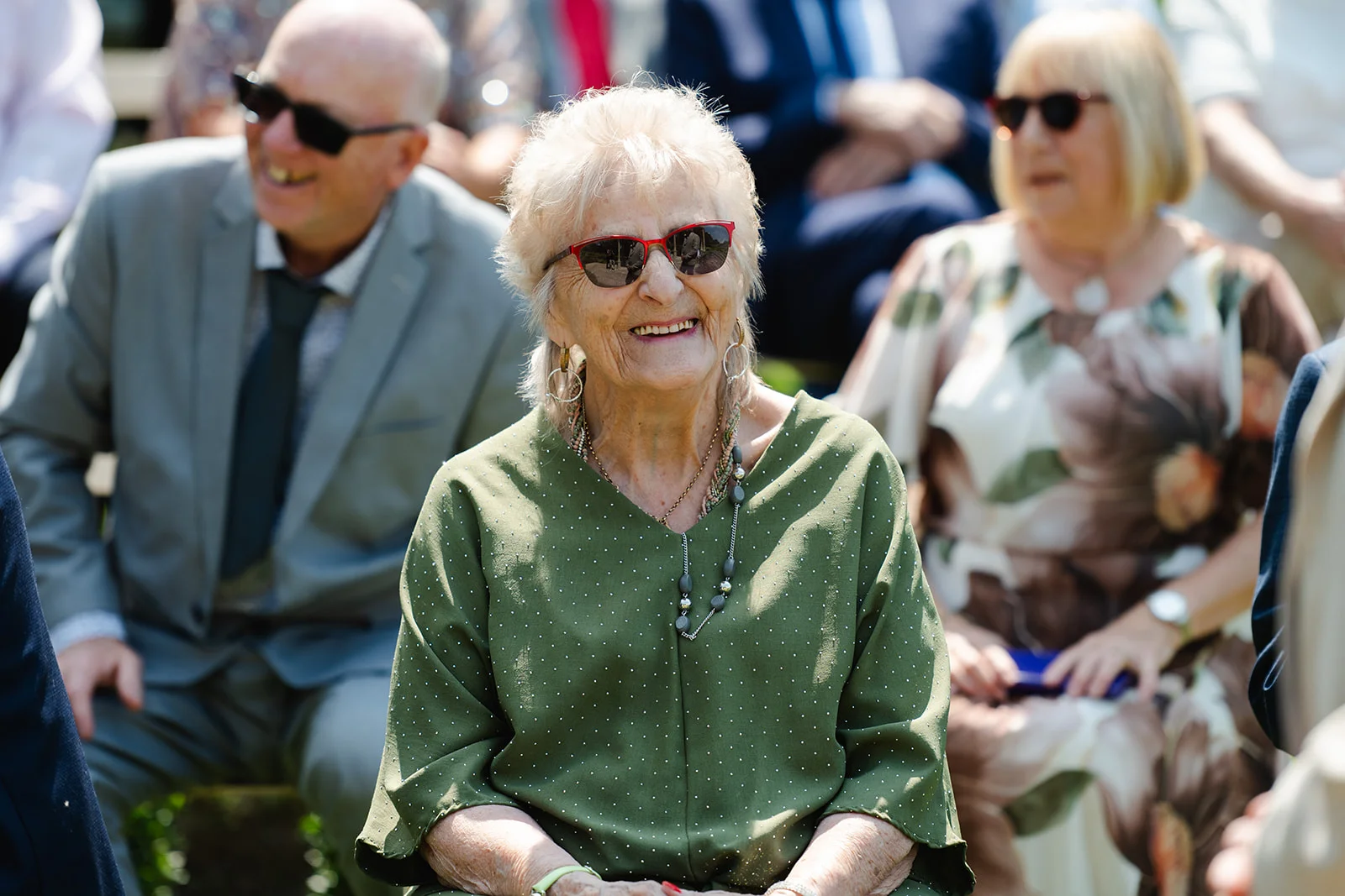 A smiling grandmother wearing sunglasses and a sage green polka dot top sits among wedding guests during a ceremony.