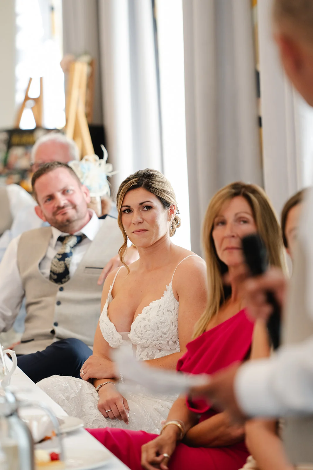A smiling bride and guests sit at the top table listening attentively to wedding speeches during the reception.