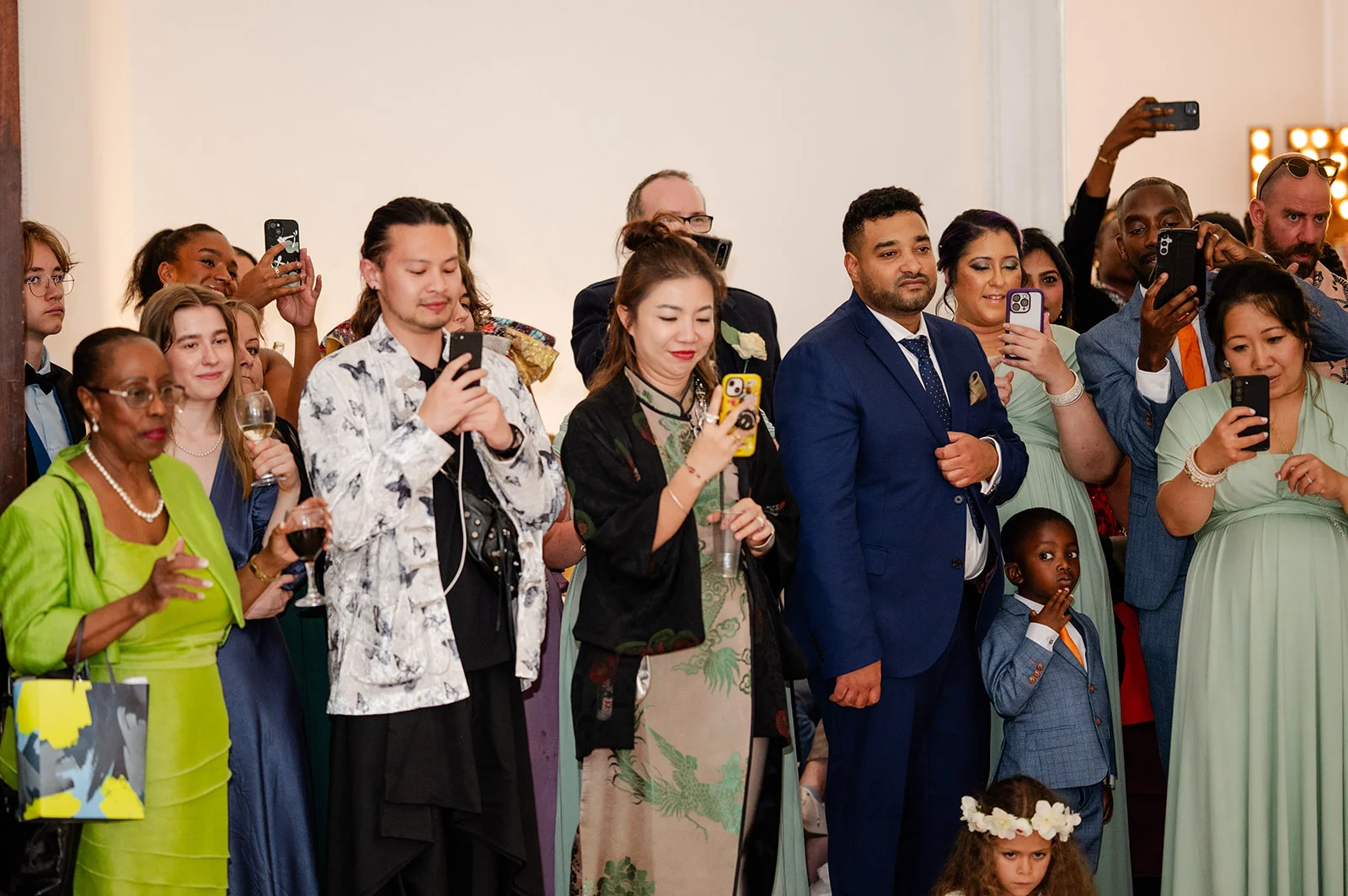 A diverse crowd of wedding guests holding up phones to record a moment, capturing engaged expressions and stylish attire.