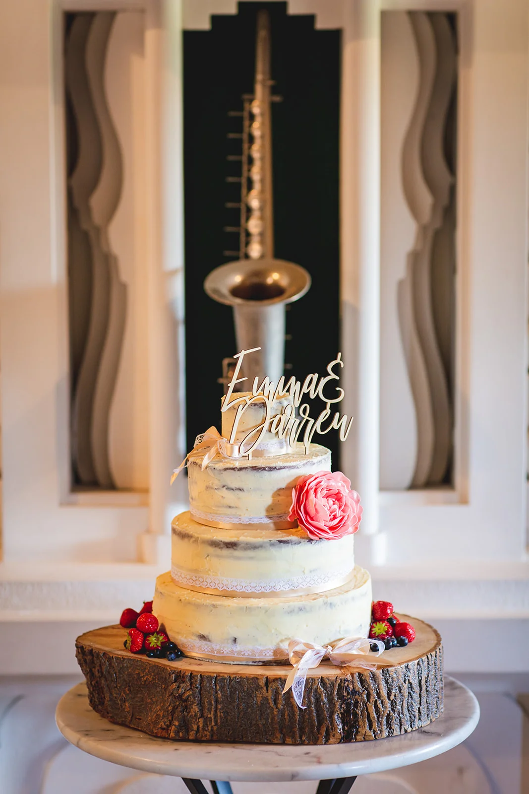 A rustic three-tier semi-naked wedding cake decorated with a large pink flower, berries, and a personalised wooden topper, resting on a tree log slice.