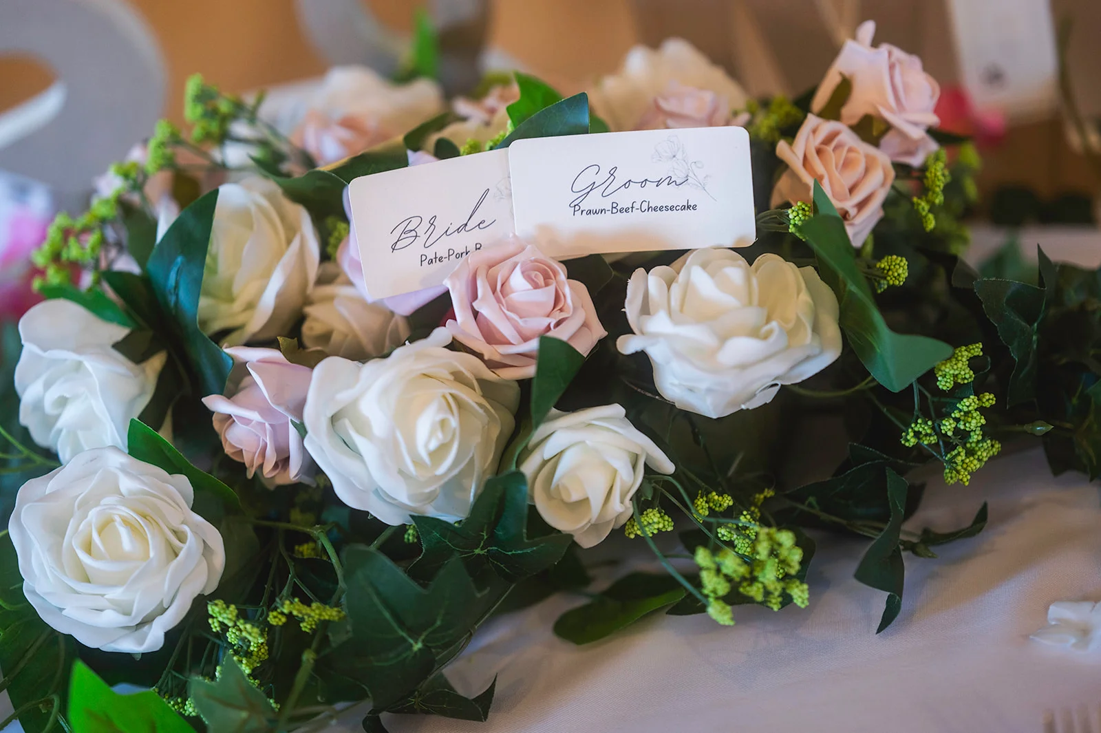 Close-up of wedding place cards labelled Bride and Groom nestled amongst an arrangement of white and pink roses with lush green foliage.