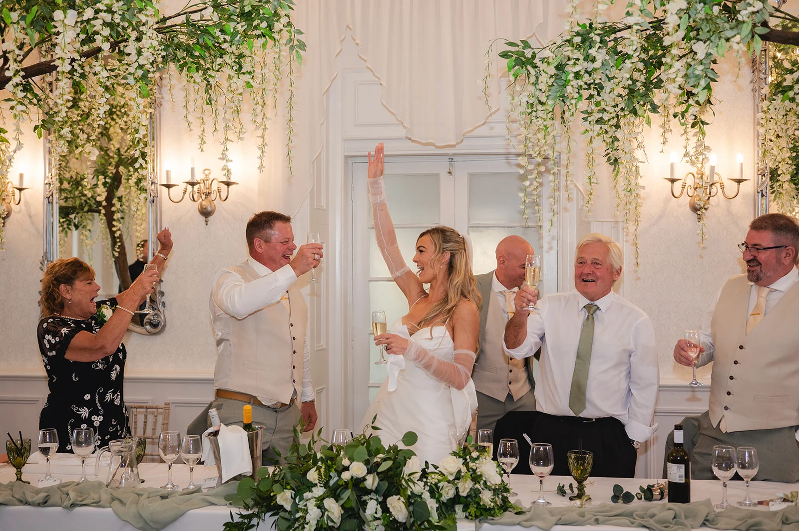 A newlywed bride raising her glass in celebration alongside smiling family and guests at the top table during wedding speeches.
