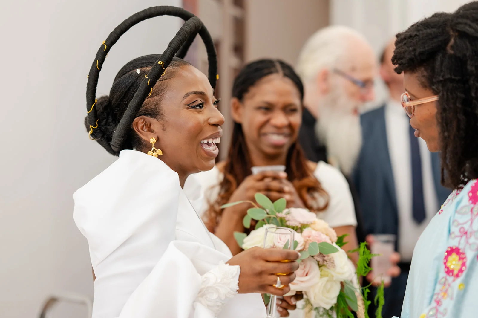 A joyful Black bride wearing a striking black halo headpiece laughs and holds a glass of champagne alongside wedding guests during the drinks reception.