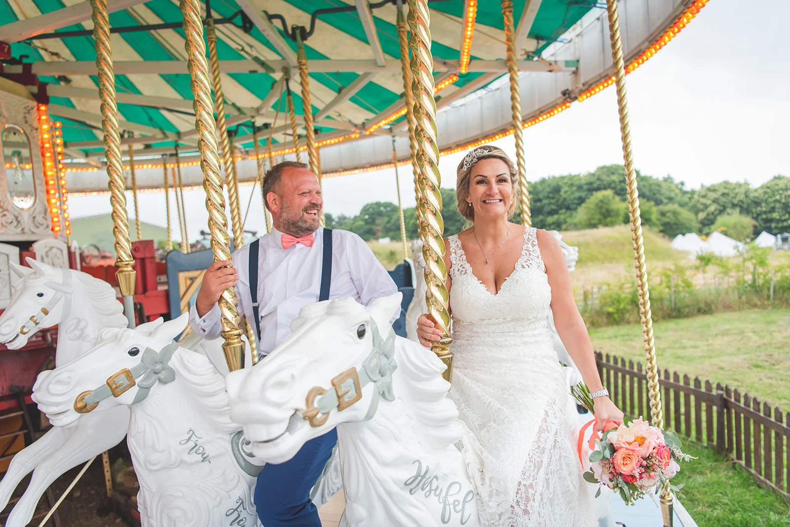 A newlywed couple happily ride a vintage carousel at an outdoor festival-themed wedding reception, with the bride holding a colourful floral bouquet.