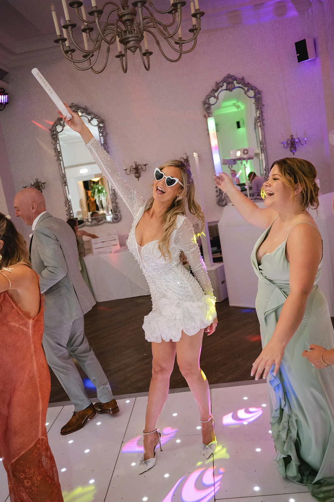 A joyful bride in a short embellished dress and heart-shaped sunglasses dances enthusiastically with guests on an illuminated dance floor during the evening reception.
