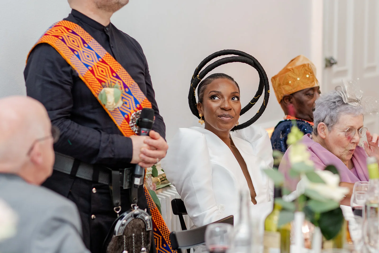 A smiling bride wearing an avant-garde circular headpiece and a structured white dress looks up intently during the wedding speeches.