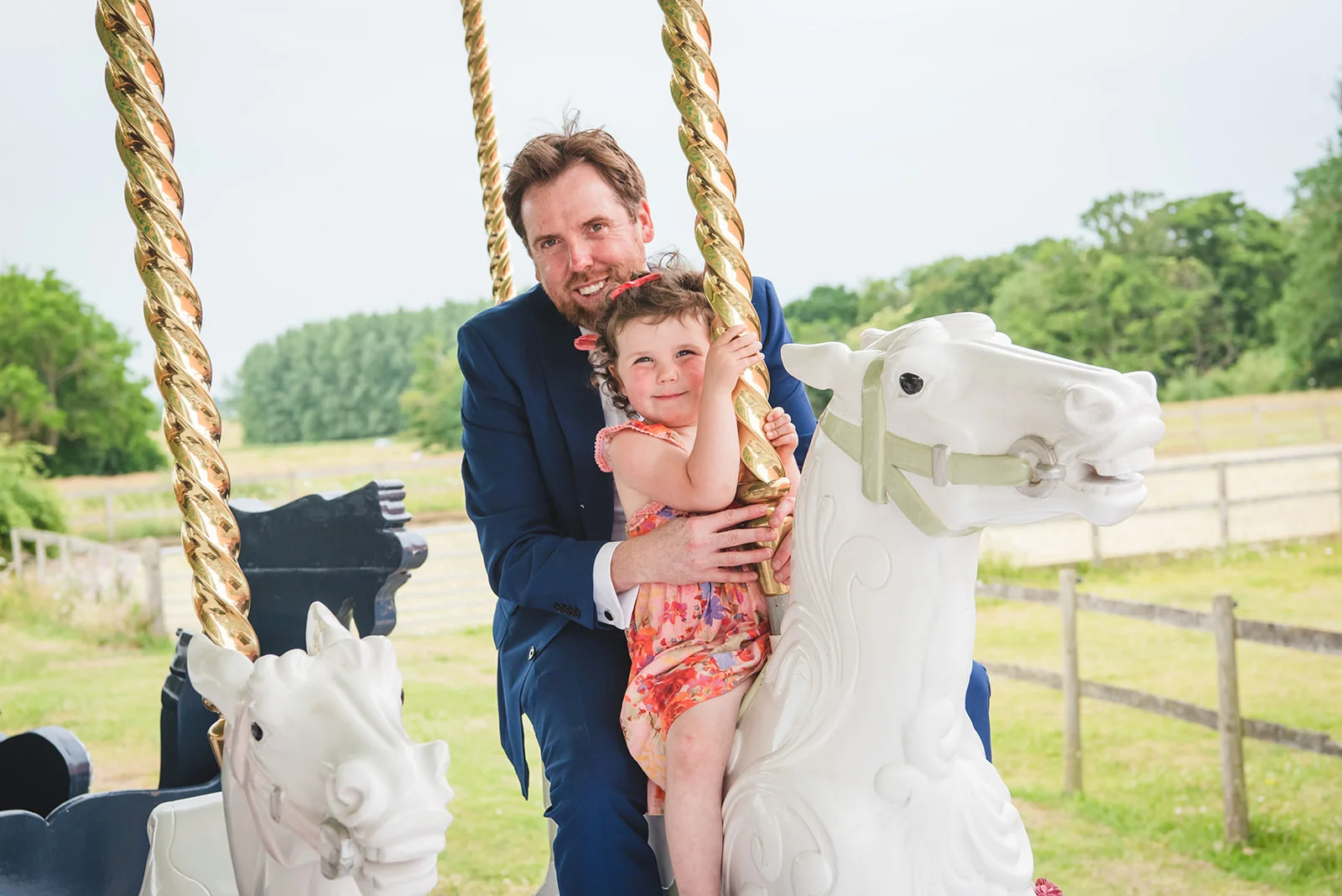 A groom and a young flower girl share a joyful moment riding on a vintage carousel during an outdoor wedding reception.