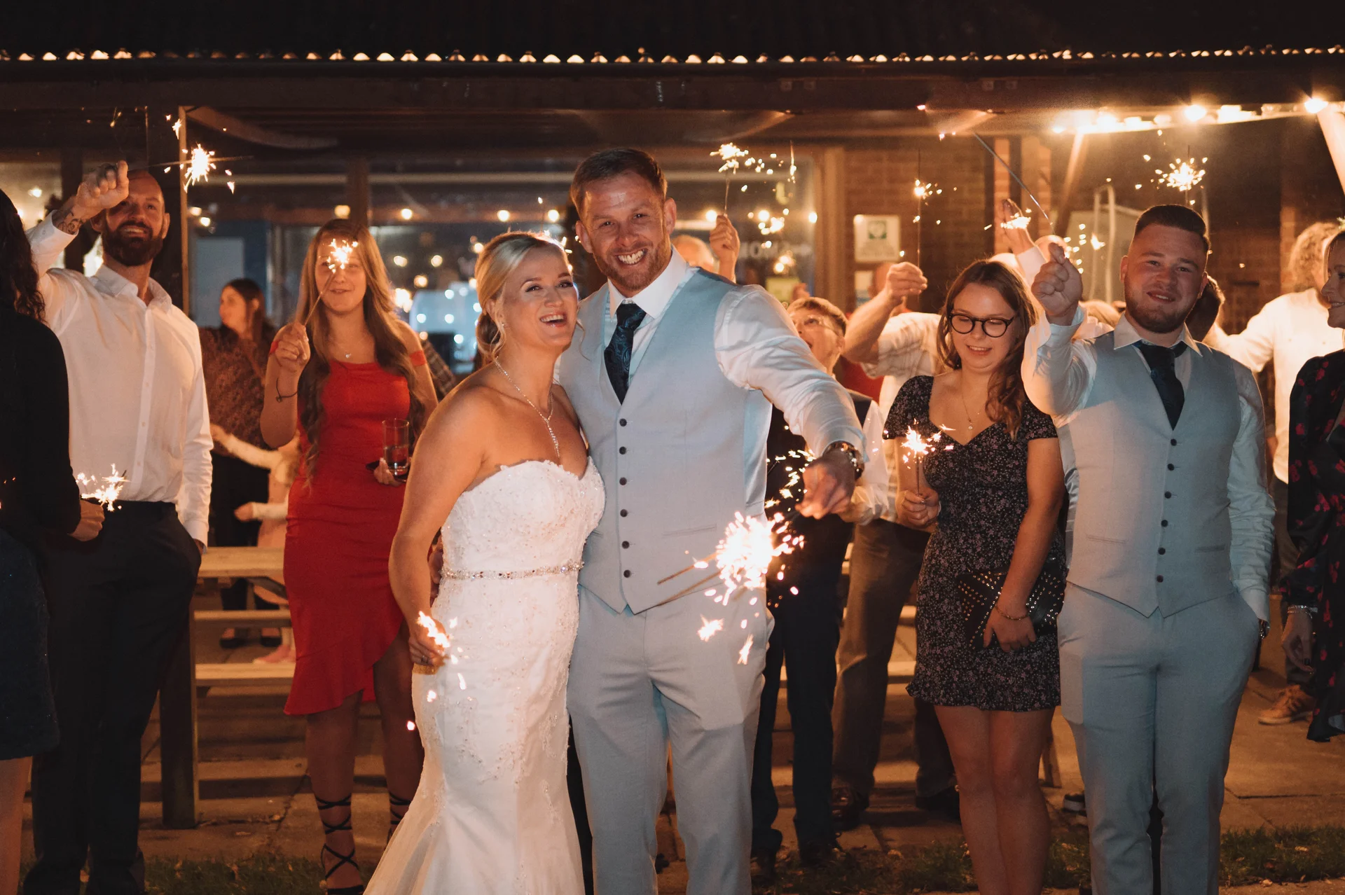 The bride and groom smile happily while holding sparklers at night, surrounded by family and friends celebrating at an evening reception.