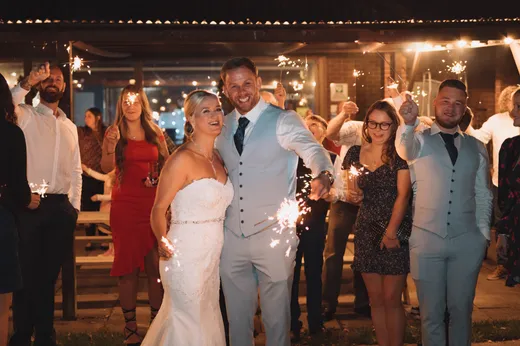 The bride and groom smile happily while holding sparklers at night, surrounded by family and friends celebrating at an evening reception.