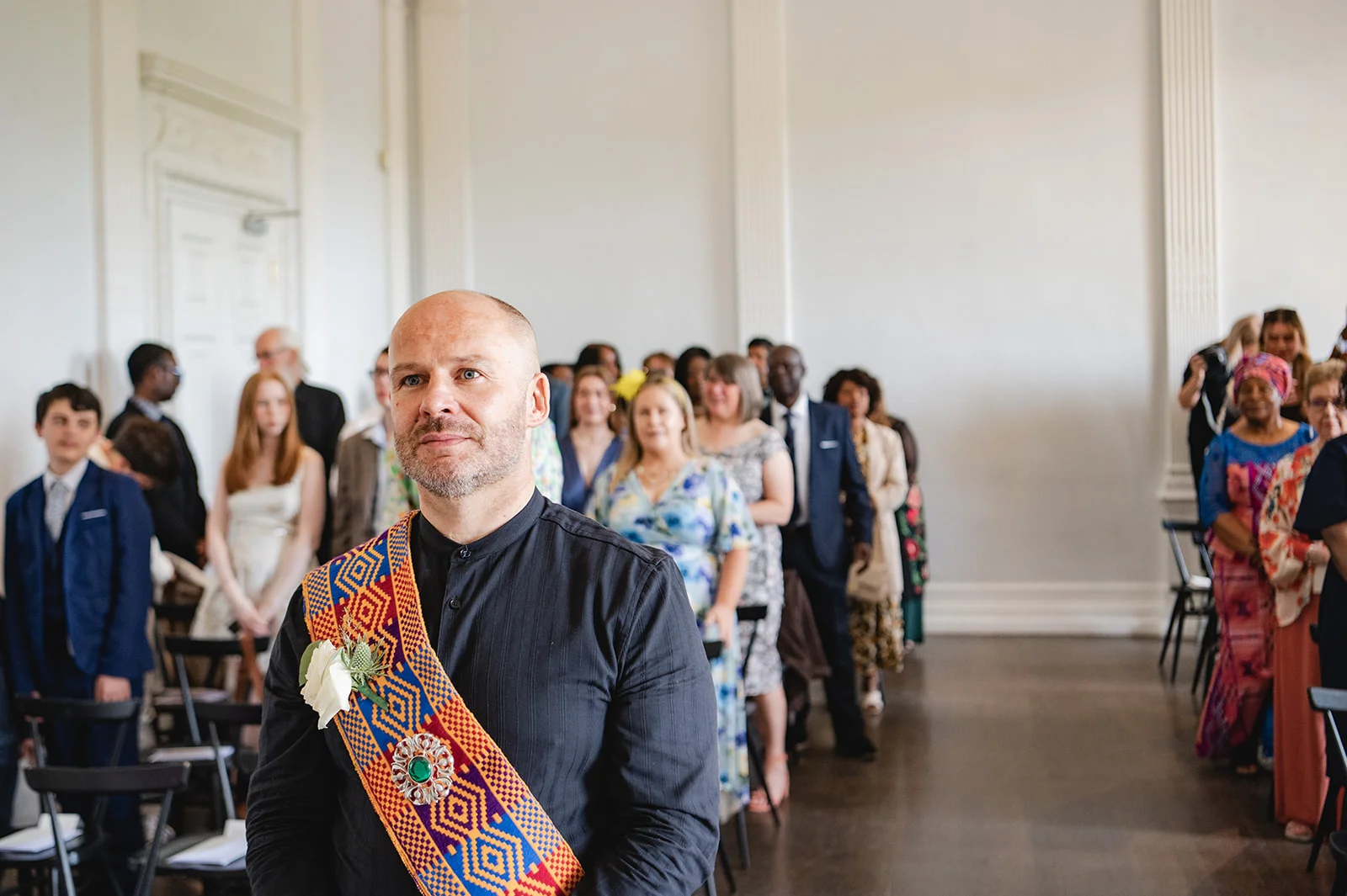 A groom wearing a patterned ceremonial sash and dark shirt stands prominently in the foreground during a wedding ceremony surrounded by seated guests.