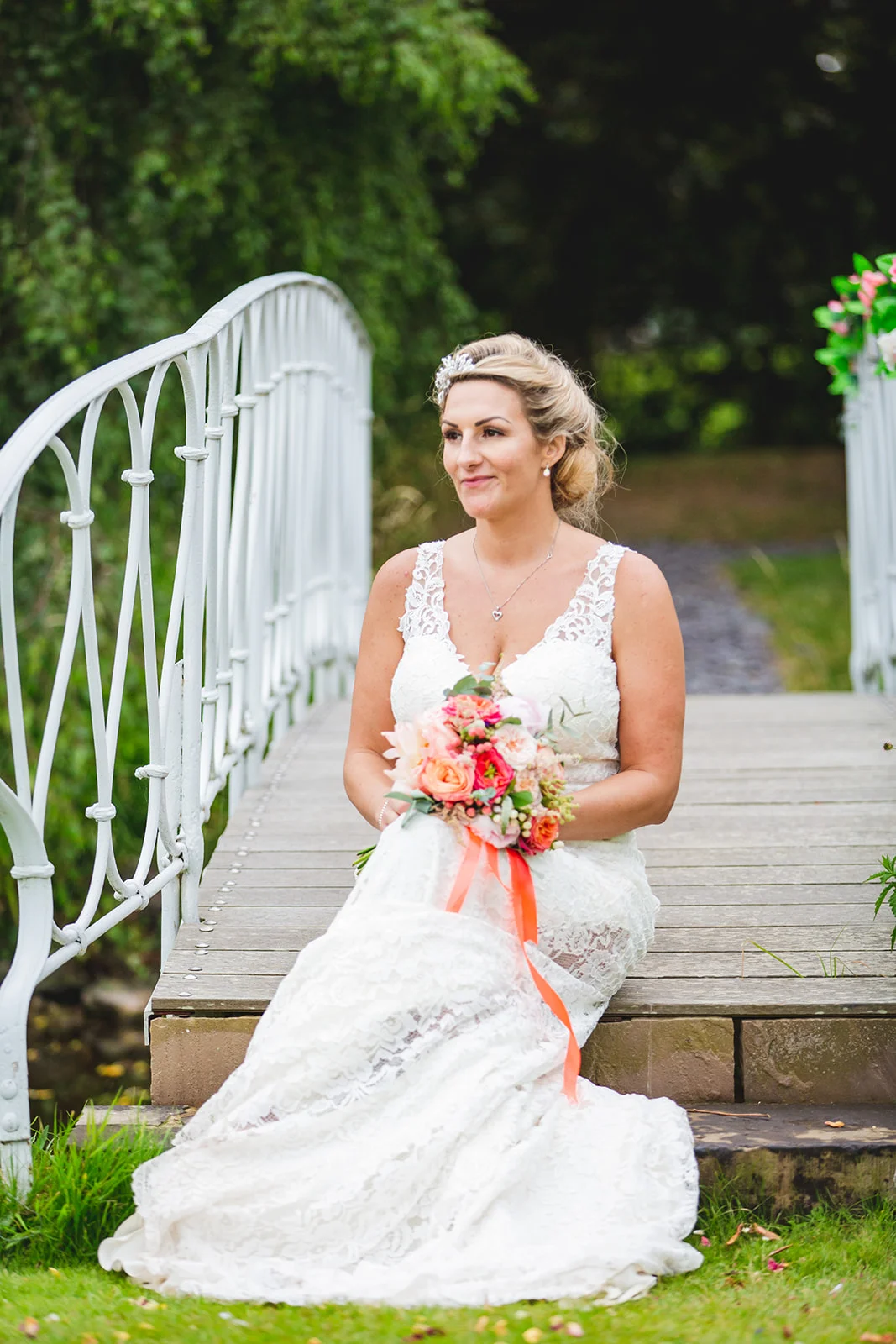 A beautiful bride sits elegantly on a wooden footbridge holding a vibrant coral and pink hand-tied bouquet.