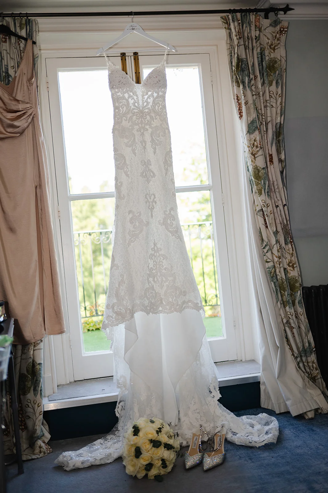An intricate lace wedding dress with spaghetti straps hangs in front of a bright window, with matching white roses and sparkly heels resting on the floor below.