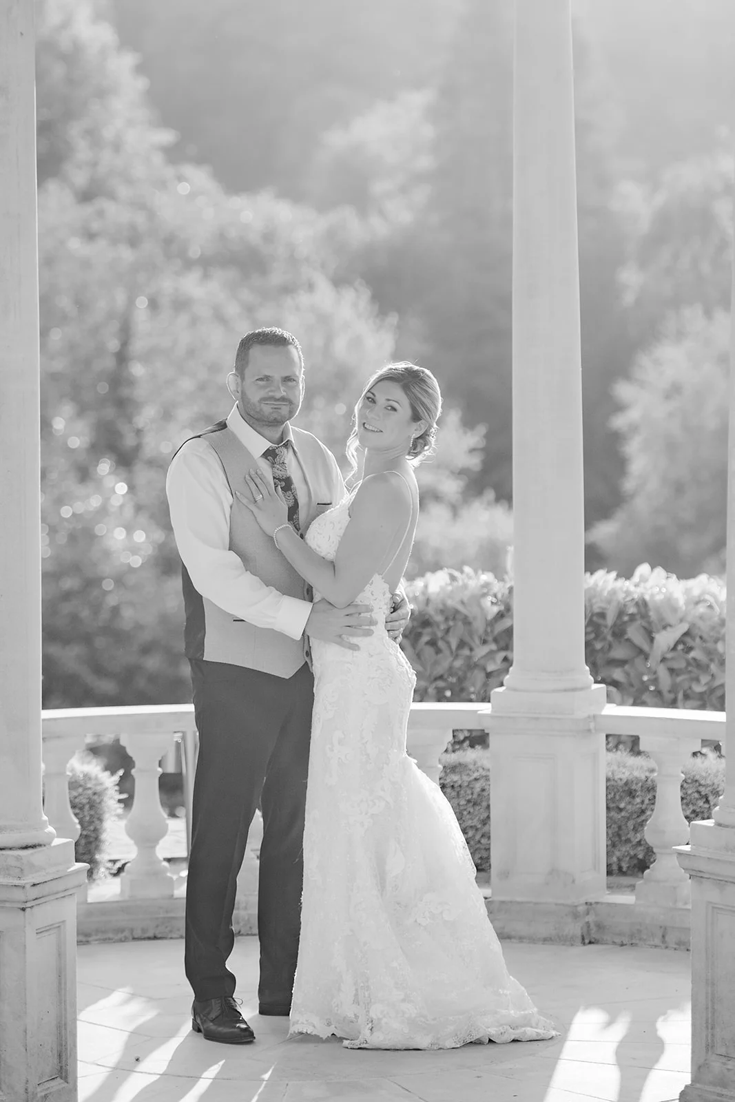 A newlywed couple poses together smiling inside a sunlit gazebo with classic pillars and balustrades during their outdoor portrait session.