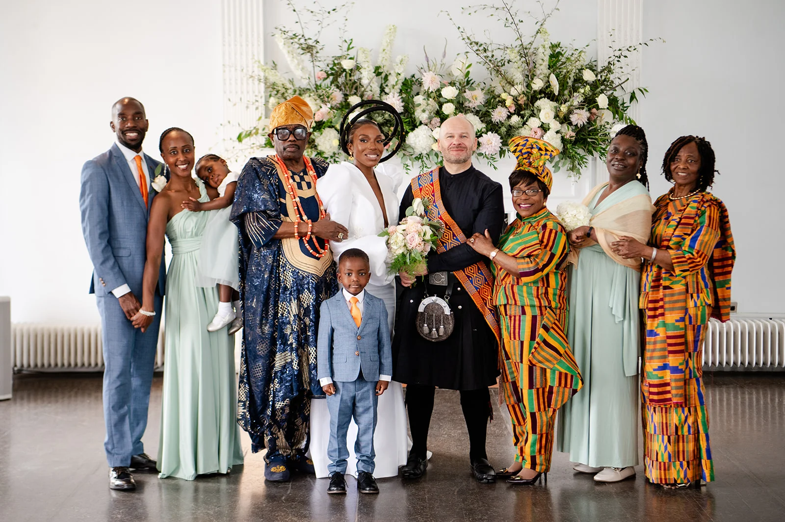 A newlywed couple posing with their multicultural wedding party and family in front of a large floral mantelpiece installation.