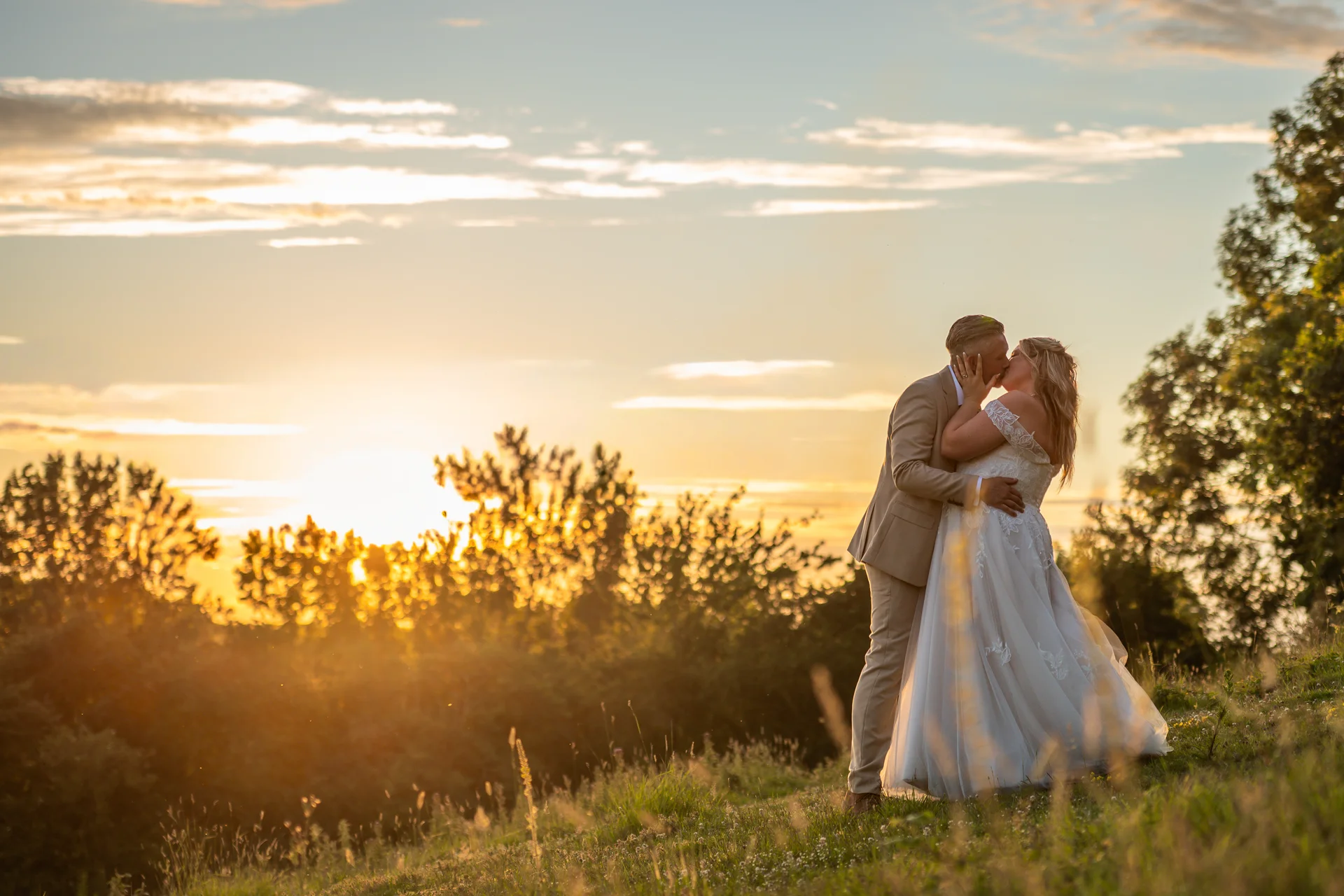Newlywed bride and groom share a tender kiss on a grassy hill during a breathtaking golden hour sunset.