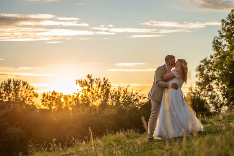 Newlywed bride and groom share a tender kiss on a grassy hill during a breathtaking golden hour sunset.