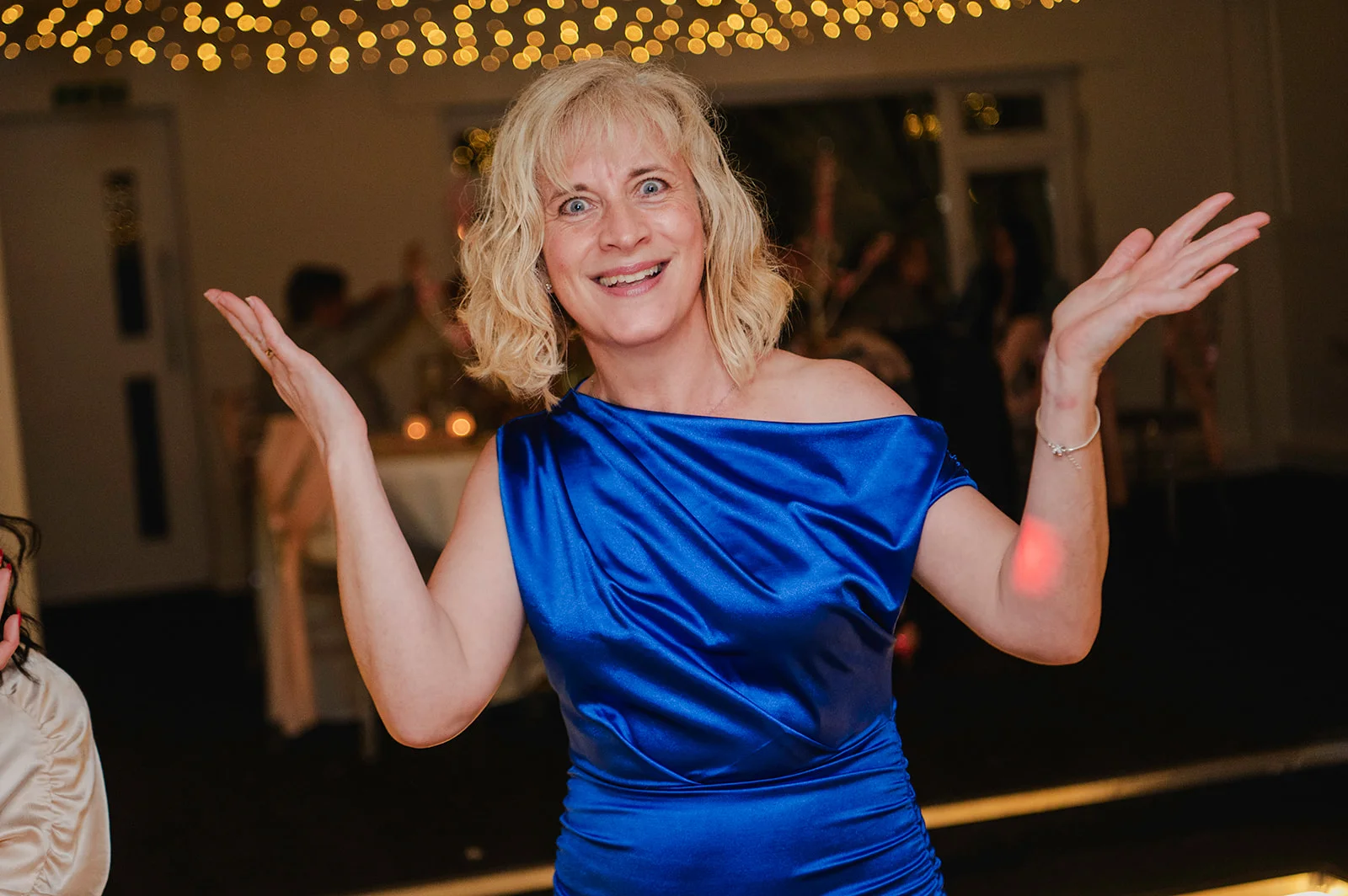 A smiling blonde guest dances enthusiastically on the reception dance floor beneath a canopy of glowing fairy lights, wearing a vibrant royal blue off-the-shoulder satin dress.