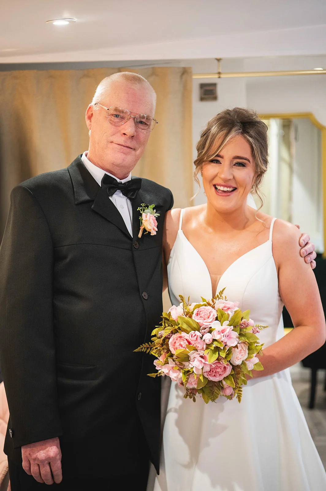 A smiling bride holding a pink floral bouquet poses alongside her father wearing a black tuxedo with a bow tie.