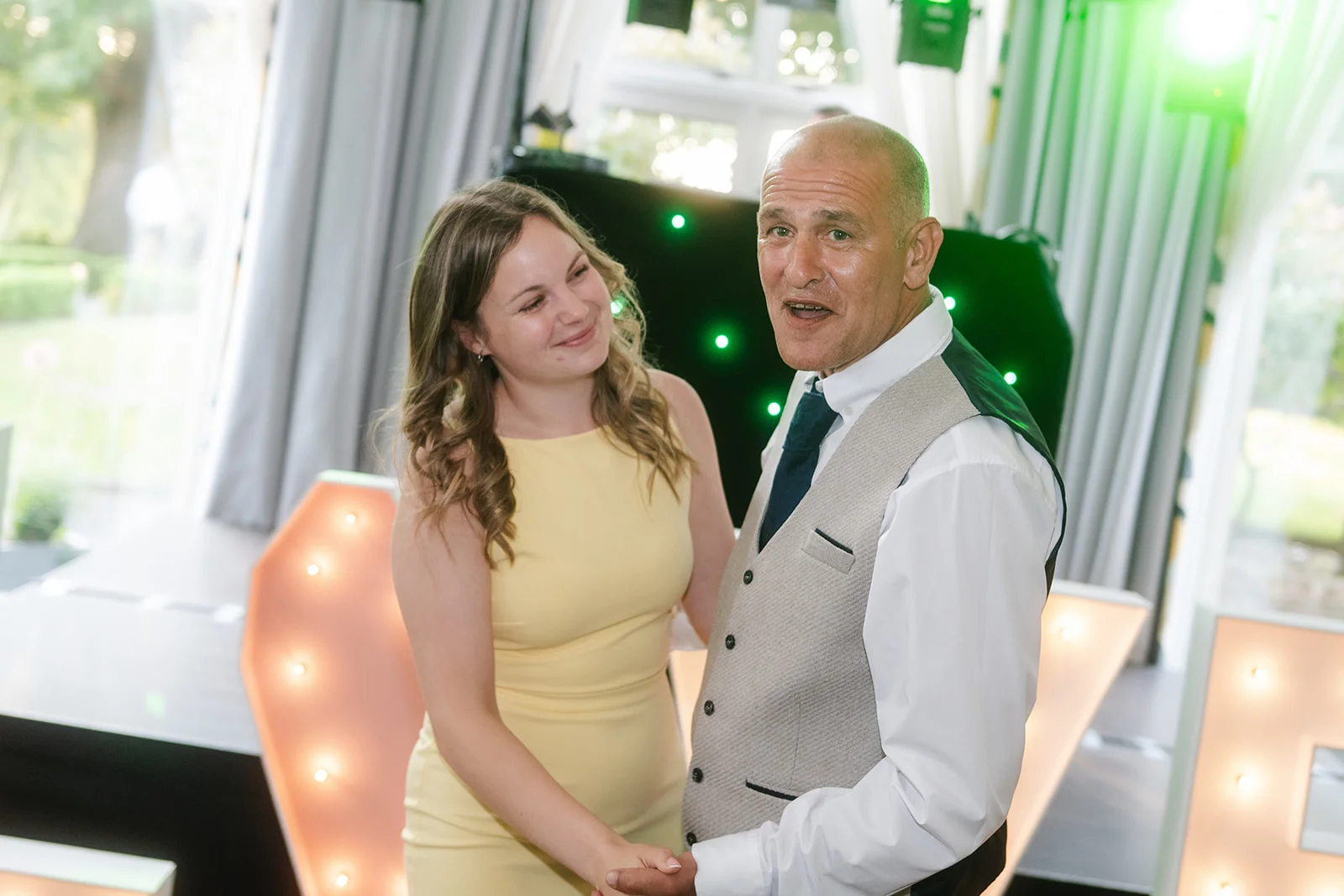 A smiling man in a grey waistcoat and a woman in a yellow dress hold hands and share a joyful moment during an evening wedding reception.
