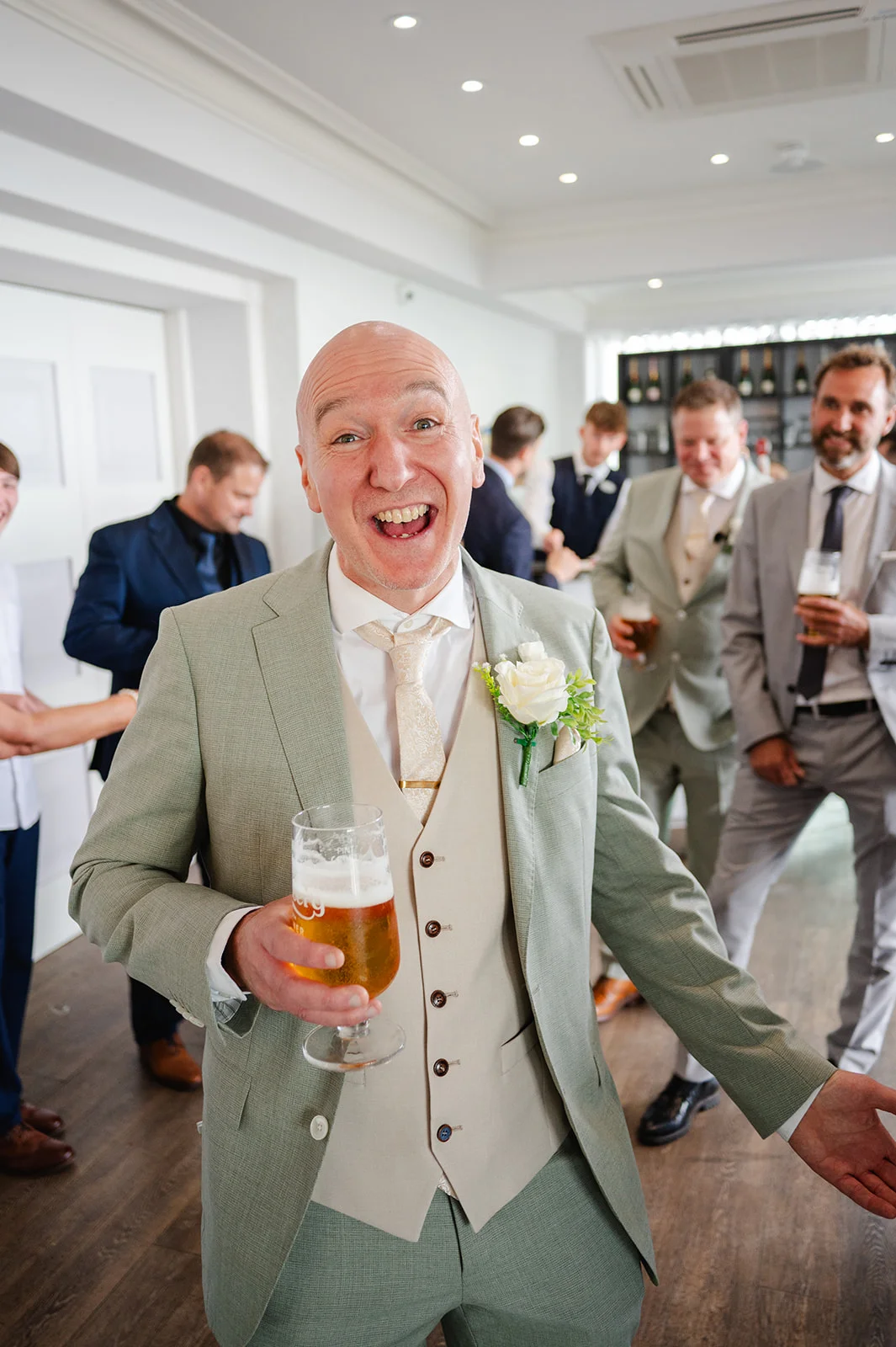 A joyful groom wearing a sage green three piece suit smiles broadly while holding a pint of beer, surrounded by wedding guests in a bright indoor venue.