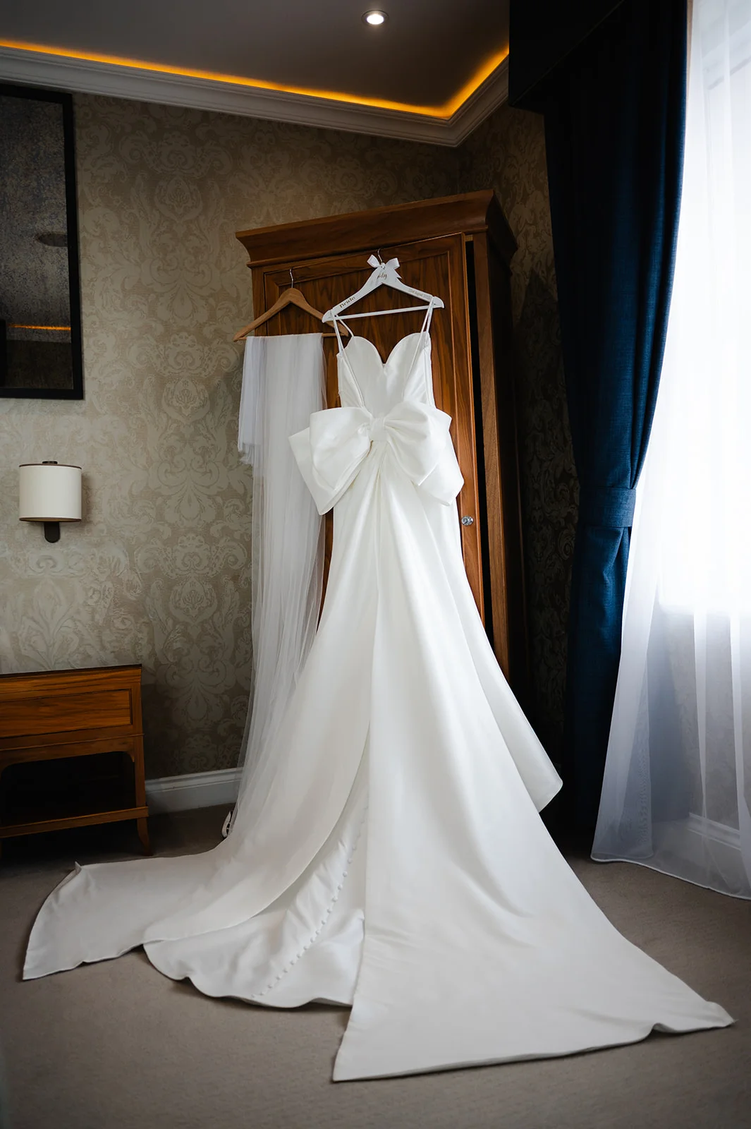 An elegant white satin wedding gown featuring a sweetheart neckline and dramatic oversized bow hangs on a wooden wardrobe in a bright hotel room.