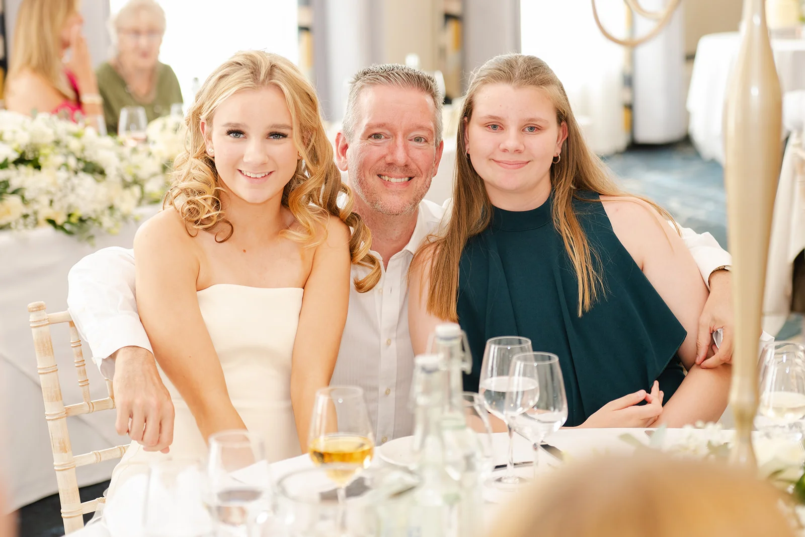 A smiling man with his arm around two young female wedding guests seated at a beautifully set reception table.
