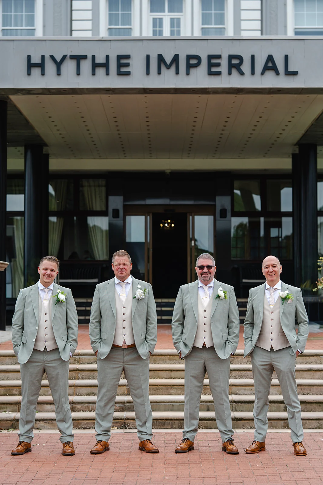 Four groomsmen stand in a line outside the Hythe Imperial hotel wearing matching sage green suits with beige waistcoats and white floral buttonholes.
