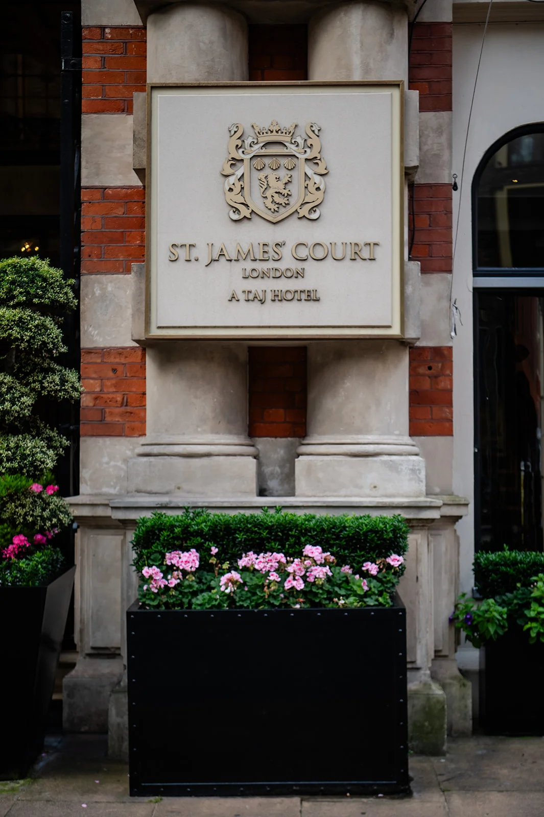 The elegant exterior entrance of St James Court, A Taj Hotel in London, featuring classic brickwork, stone architectural columns, and sophisticated signage above floral planters.