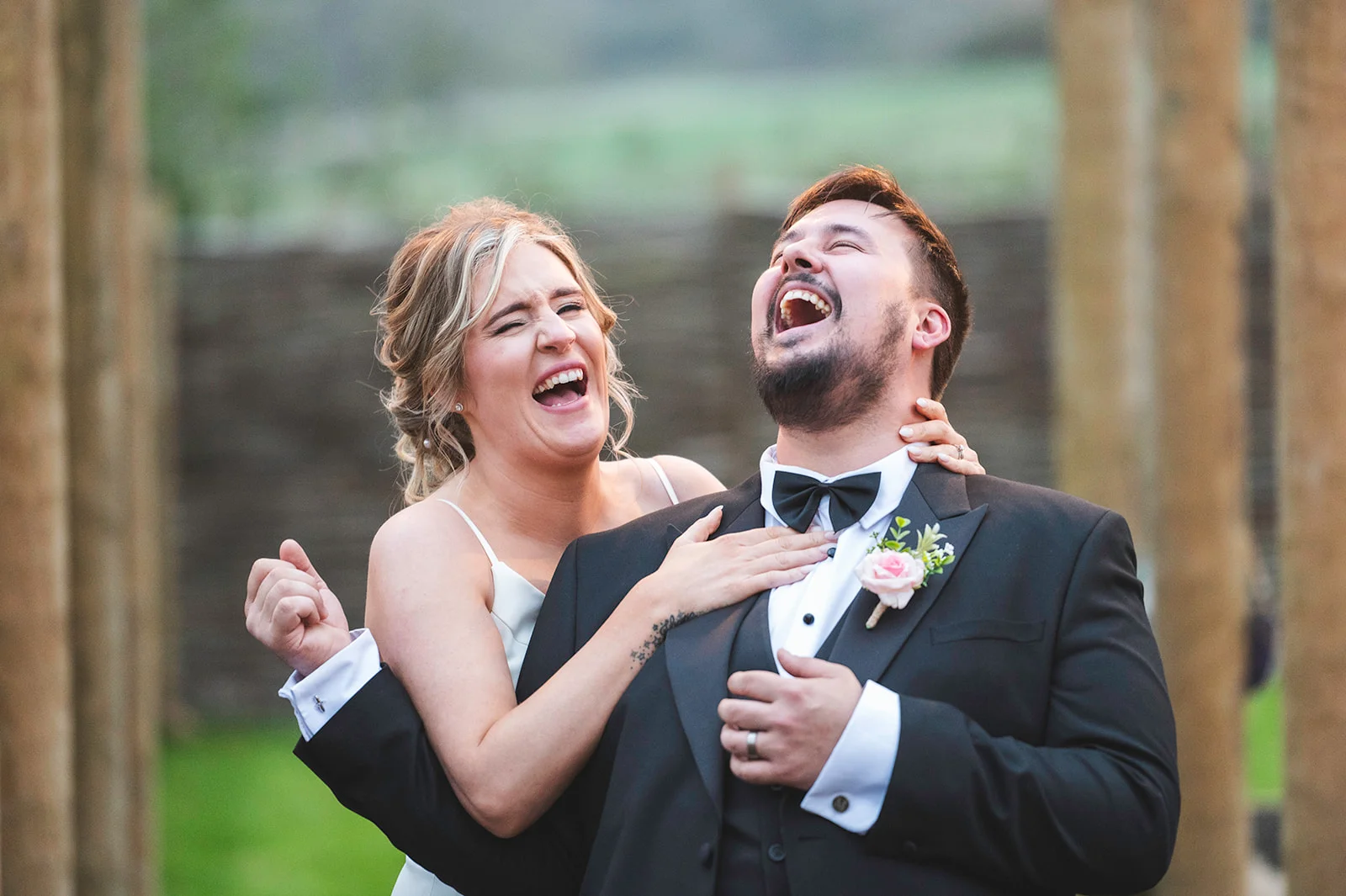 A newlywed couple share a joyful, laughing embrace outdoors in golden backlighting, with the groom wearing a classic black tuxedo and the bride in a simple white gown.