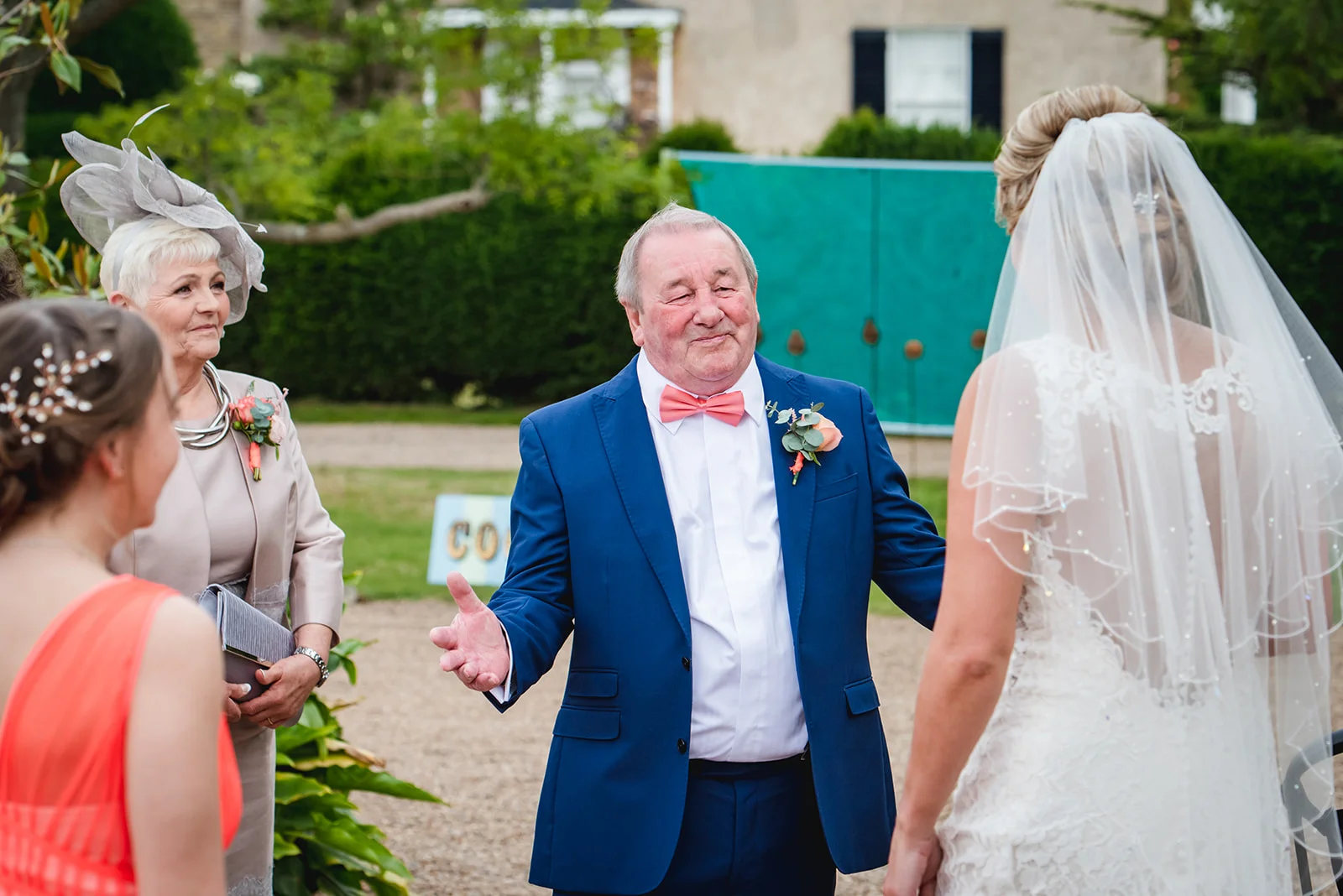 An emotional father in a blue suit and pink bow tie gestures warmly toward the bride in her lace gown and veil, while family members look on outdoors.