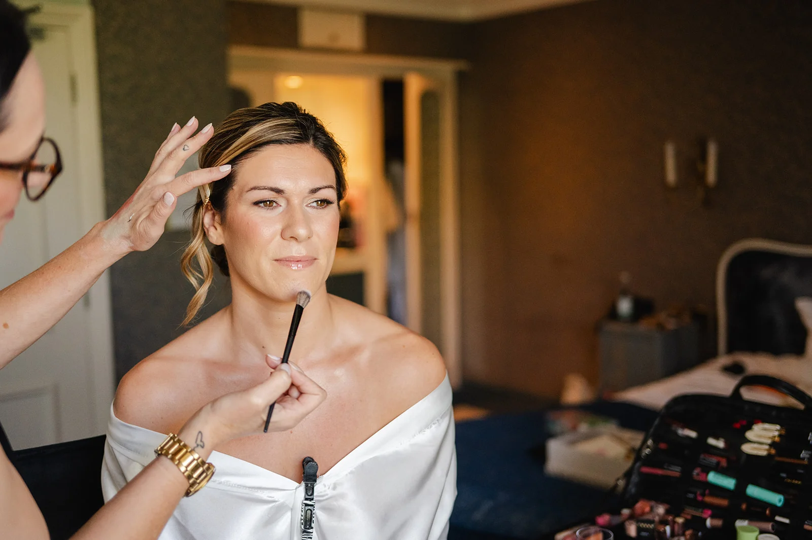 A professional makeup artist applies finishing touches to a bride wearing a white getting ready robe in a softly lit bedroom.