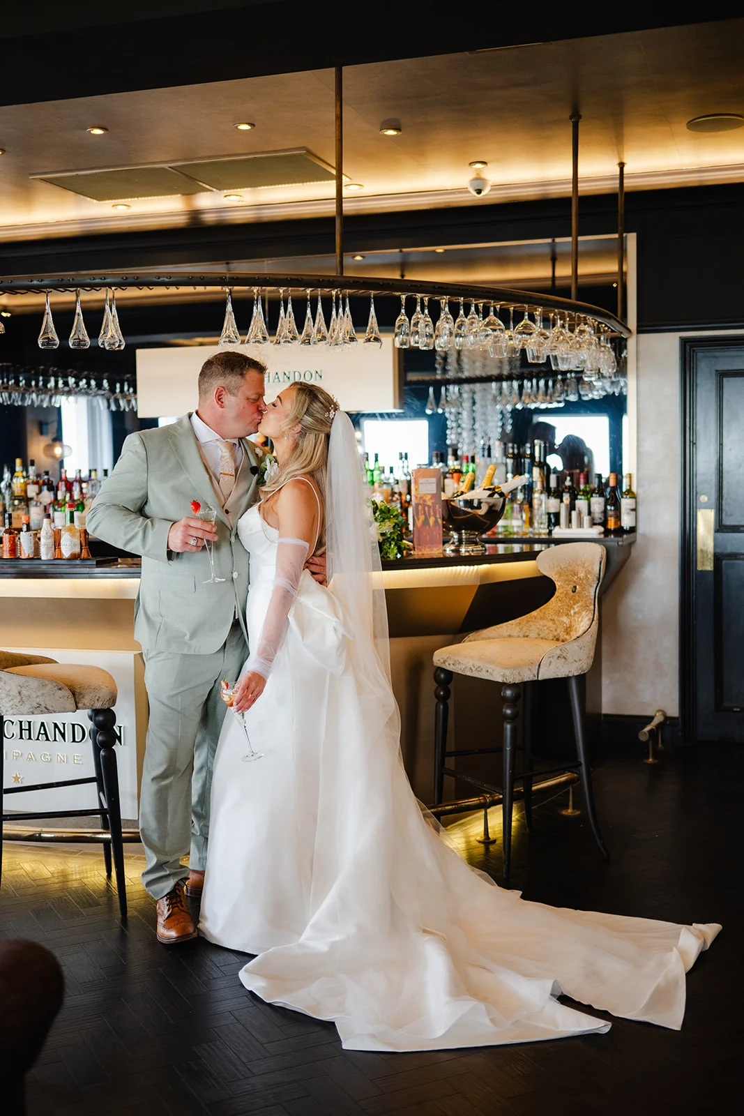 A bride and groom share a kiss inside a stylish bar with hanging glassware, while holding cocktail glasses near the counter.