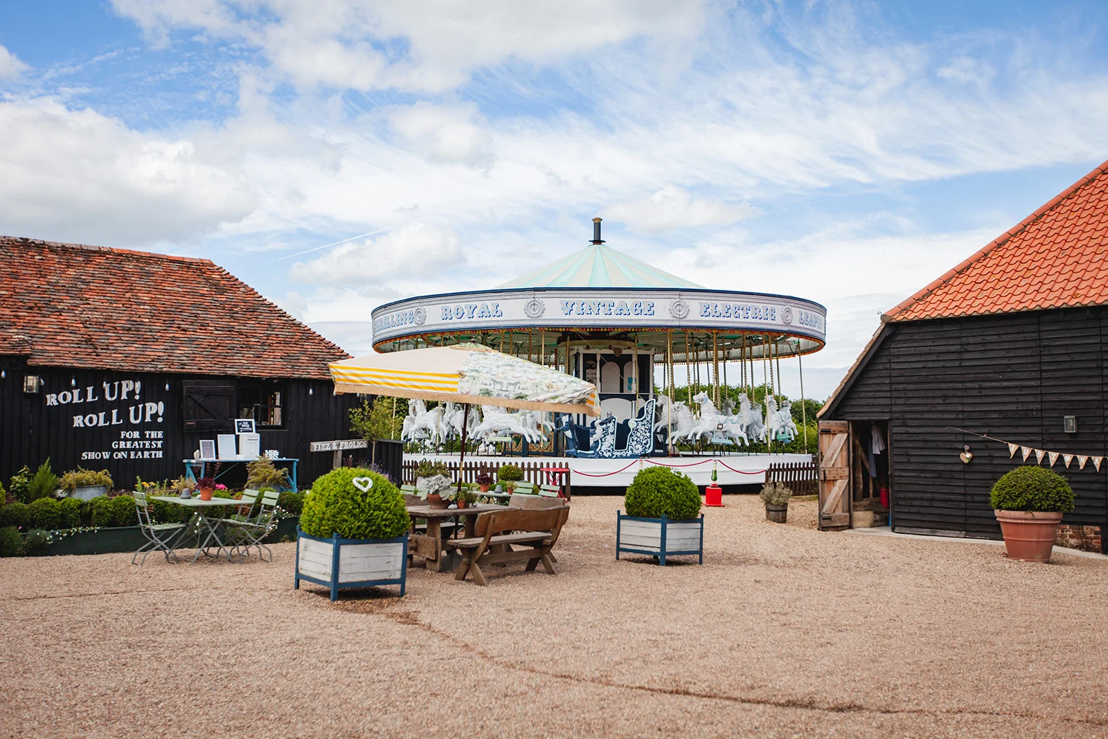 A vintage fairground carousel sits in the courtyard of a rustic barn venue under a bright blue sky on a sunny wedding day.