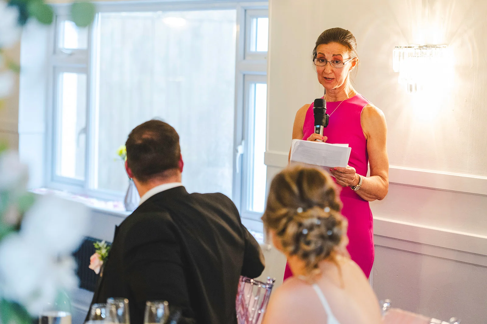 A mother of the bride delivers a heartfelt speech holding a microphone and notes, while the couple listens attentively from their table.