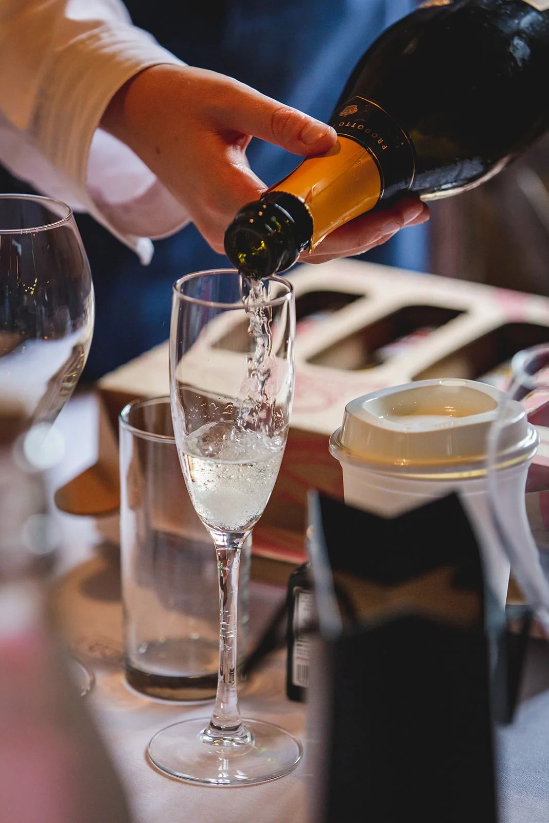 A close-up shot captures sparkling wine being poured from a bottle into a tall flute glass during morning bridal preparations.